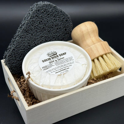 Solid dish soap in a ramekin container, with a bamboo brush and soap sponge in a small wooden box. It's displayed on a dark background.