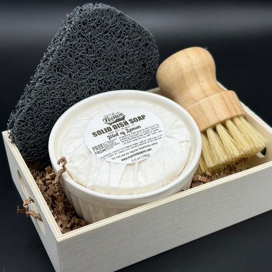 Solid dish soap in a ramekin container, with a bamboo brush and soap sponge in a small wooden box. It's displayed on a dark background.