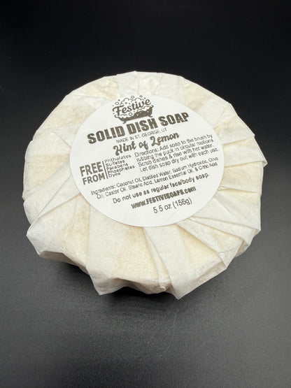 Round bar of solid dish soap, wrapped with a white paper and a product label on a dark background. It contains a lemon fragrance and lists the ingredients.