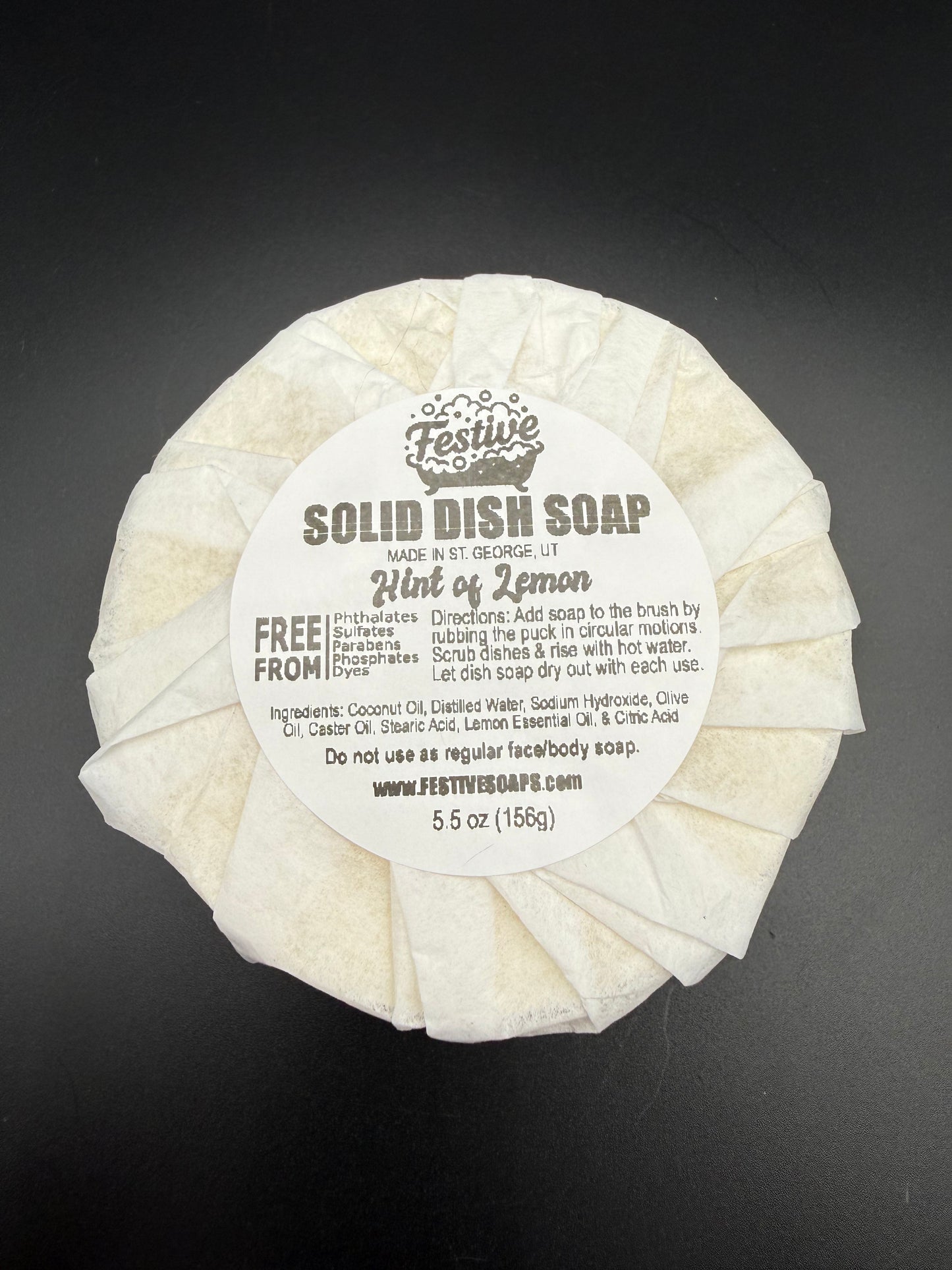 Round bar of solid dish soap, wrapped with a white paper and a product label on a dark background. It contains a lemon fragrance and lists the ingredients.