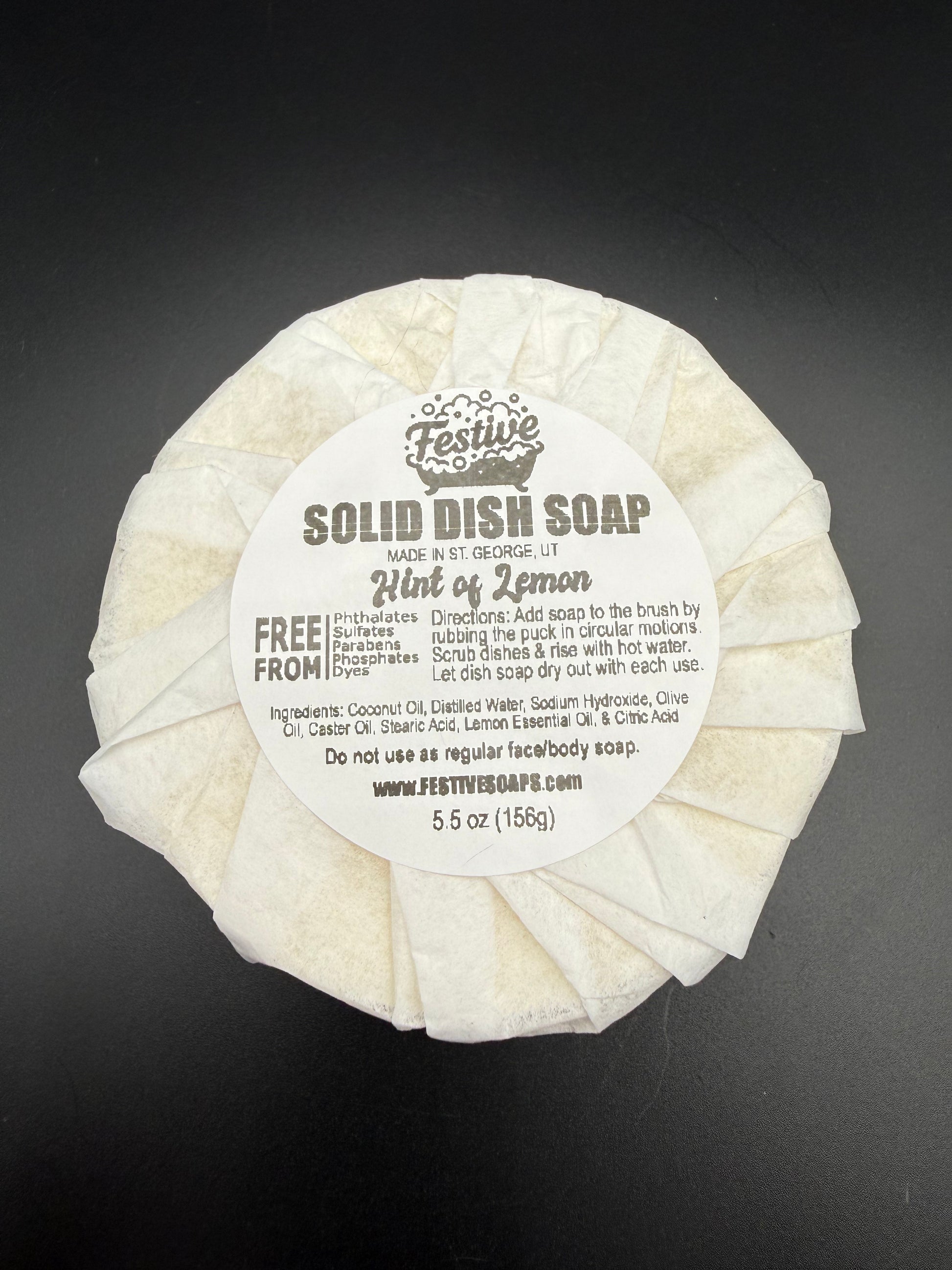 Round bar of solid dish soap, wrapped with a white paper and a product label on a dark background. It contains a lemon fragrance and lists the ingredients.