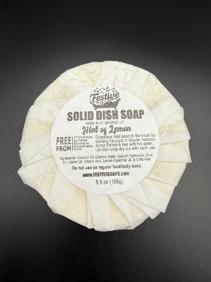 Round bar of solid dish soap, wrapped with a white paper and a product label on a dark background. It contains a lemon fragrance and lists the ingredients.